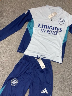 Arsenal träningsset från Adidas - Snyggt Arsenal träningsset med långärmad tröja och byxor i blått och ljusblått. Tröjan har dragkedja vid halsen och tryckt Arsenal-logga samt sponsortryck. Byxorna har resår i midjan och Adidas-logga. Perfekt för fotbollsträning eller chill.