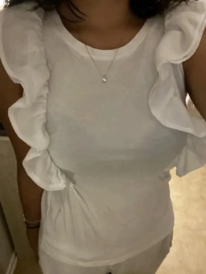 Vit volangtopp med ärmlös design - Supersöt vit topp med volangdetaljer längs axlarna och ärmlös design. Perfekt för dig som gillar en feminin och fräsch stil. Tillverkad i mjukt bomullstyg som känns skönt mot huden. Rund halsringning och figurnära passform.