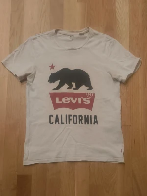 Vintage Levi's t-shirt - Vit t-shirt från Levi's med tryck av en svart björn, röd stjärna och texten 'Levi's California' på framsidan. Klassisk passform med rund hals och korta ärmar. Perfekt för en avslappnad stil.