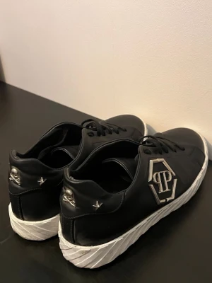 Philipp Plein svarta sneakers med dödskalle - Svarta sneakers i läder från Philipp Plein med vita, mönstrade sulor. Sidorna pryds av en stor metallogo och bak finns dödskalle-detaljer samt nitar i silver. Snygg och edgy design som sticker ut.