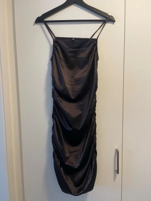 Svart satin kortklänning med smala axelband - Snygg svart kortklänning i satin med smala axelband och draperad effekt längs sidorna. 