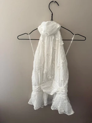 Vit volangig ärmlös blus - Supersöt vit blus med volangkant och tunna axelband. Topp i lätt och luftig bomull med små broderade prickar och blommor. Perfekt för varma dagar och ger en romantisk vibe till din outfit.