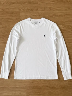 Vit långärmad topp från Ralph Lauren - Klassisk vit långärmad topp från Ralph Lauren med rund halsringning och ikonisk broderad logga på bröstet. Tillverkad i mjuk bomull som känns skön mot huden. Perfekt för en clean och stilren look.