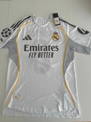 Real Madrid matchtröja Adidas player version - Snygg vit Real Madrid matchtröja från Adidas med klubbmärke på bröstet, gråa detaljer och gula linjer längs sidorna. Tröjan har trycket 'Emirates Fly Better' på framsidan och Champions League-märke på ärmen. Tillverkad i lätt och ventilerande material.
