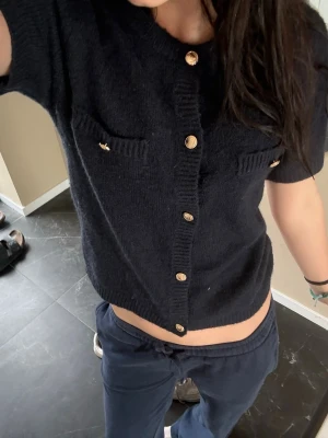 Mörkblå stickad kortärmad topp - Mörkblå stickad topp med korta ärmar och snygga guldknappar framtill. Två dekorativa bröstfickor med knappdetaljer ger en cool touch. Perfekt att styla med både jeans och kjol. Storlek 179 från Gina