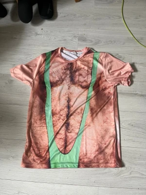 T-shirt med kroppstryck och gröna band - Rolig kortärmad t-shirt i syntetmaterial med tryck som föreställer en hårig överkropp och gröna baddräktsband. Färgerna är rosa, brun och grön. Passformen är loose och t-shirten har rund hals.