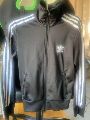 Svart Adidas hoodie med vita ränder - Säljer en svart Adidas hoodie med dragkedja och klassiska vita ränder längs ärmarna. Framtill finns Adidas logga på bröstet och en stor vit trefoil-logga på ryggen. Mjuk och sportig modell i syntetmaterial, perfekt för en streetwear-look.