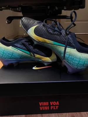 Nike Mercurial Vini Fly fotbollsskor - Snygga Nike Mercurial fotbollsskor med svart ovandel och färgskiftande sidor i gult, grönt och rosa. Skorna har texten 'VINI VOA VINI FLY' tryckt på sidan och en stor Swoosh-logga. Lätta material och låg profil för maxad speed på planen.