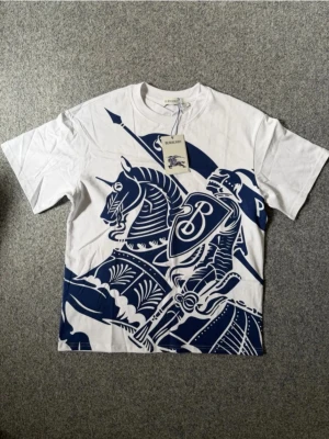 Vit Burberry t-shirt - Säljer en vit t-shirt från Burberry med ett stort blått tryck av en riddare på häst över hela framsidan. T-shirten har rund halsringning och korta ärmar. Perfekt statement-plagg för dig som gillar unika designer.