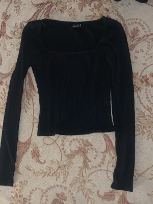 Svart långärmad topp från Gina Tricot - Snygg svart långärmad topp från Gina Tricot med fyrkantig ringning och ribbad struktur. Perfekt att styla med jeans eller kjol för en trendig look. Toppens material känns mjukt och följsamt mot kroppen.
