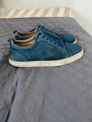 Blåa sneakers med nitar från Louboutin - Unika blåa sneakers i mocka från Louboutin med coola nitar på tån och klassisk röd sula. Skorna har blå snören och en vit sula som ger en snygg kontrast. Perfekta för dig som vill sticka ut med din stil. Men det går att ta in de hos en skomakare så kommer det bli nästan som nya 