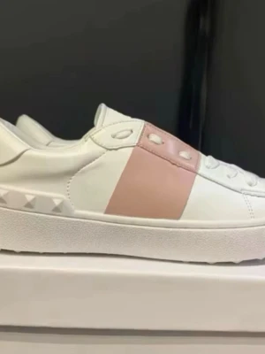 Vita sneakers med rosa detalj - Snygga vita sneakers från Valentino med en bred rosa läderdetalj över sidan och coola nitar på hälen. Skorna har en klassisk låg profil och är tillverkade i slätt läder med vita snören. Perfekta för dig som vill ha en stilren men ändå unik look.