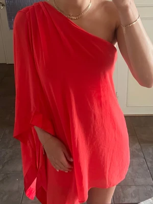 Röd oneshoulder enaxlad miniklänning kortklänning storlek S/36 - Säljer en röd kortklänning med oneshoulder-design. Klänningen har en lös passform och är ärmlös på ena sidan medan den andra sidan har ett draperat tyg som ger en asymmetrisk look. Perfekt för dig som vill sticka ut med en enkel men snygg stil. Finns lite dragen tråd i tyllen, syns dock knappt när den bärs.