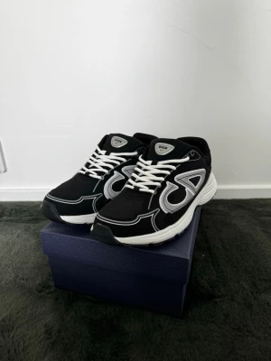 Dior B30 svarta sneakers - Svarta Dior B30 sneakers med vit sula och vita skosnören. Skorna har stora reflekterande silverdetaljer på sidorna och meshpaneler för extra andningsförmåga. Snygg och sportig design med tydlig Dior-logga framtill och på sidan.