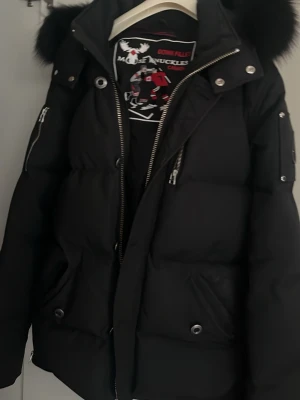 Svart dunjacka från Moose Knuckles - (Äkta med kvitto) Snygg svart dunjacka från Moose Knuckles med pälskantad huva, silverfärgade dragkedjor och tryckknappar. Jackan har flera fickor och en patch med logga på ärmen. Perfekt för kalla vinterdagar och riktigt varm. Kan gå ner mycke i pris vid snabb affär