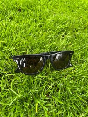 Svarta Ray-Ban Wayfarer solglasögon - Klassiska svarta Ray-Ban Wayfarer solglasögon med lite brunaktig lins och svart båge och ikonisk fyrkantig form. Snygg och tidlös design med Ray-Ban-logga på bågen. Perfekta för att lyfta din stil under soliga dagar.