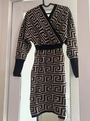 Beige och svart omlottklänning - Snygg kort omlottklänning i beige och svart med grafiskt mönster. Klänningen har långa ärmar, markerad midja och breda muddar. Perfekt för dig som gillar unika detaljer och trendig design.
