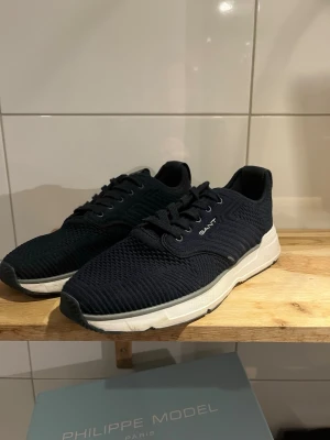 Marinblå sneakers från Gant - Snygga marinblå sneakers från Gant med ovandel i stickad textil och vita sulor. Skorna har klassisk låg profil, svarta skosnören och diskret Gant-logga på sidan. Perfekta för en stilren och sportig look.