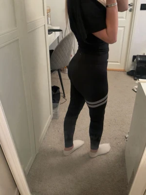 Svarta tights med vita ränder - Snygga svarta leggings från Stronger med två vita ränder runt ena låret. Hög midja och tight passform som framhäver figuren. Perfekta för träning eller chill. Materialet är stretchigt och bekvämt.