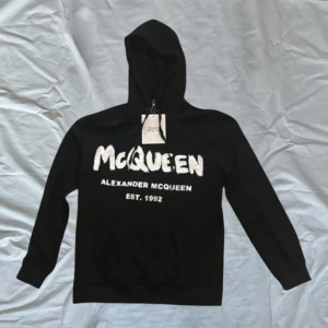 Svart hoodie från Alexander McQueen - Säljer en snygg svart hoodie från Alexander McQueen med stort vitt tryck på framsidan. Den är i nyskick och har en klassisk passform med långa ärmar och en bekväm huva. Den är väldigt bra skick nypris runt 8000 M/S pris är hugget i sten 