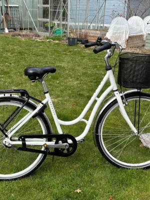 Vit Monark Original  - Säljer en vit klassisk damcykel med lågt insteg, svart pakethållare och en praktisk svart cykelkorg fram. Cykeln har stänkskärmar, kedjeskydd och bekvämt sadel. Perfekt för stadskörning och vardagspendling. Båda nycklar medföljer original låset. 