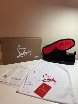 Svarta sneakers från Christian Louboutin - Exklusiva svarta sneakers från Christian Louboutin med ikonisk röd sula och diskret logga. Skorna har en klassisk låg design och är tillverkade i lyxigt material med snygga detaljer. Perfekt för dig som vill sticka ut med stil.