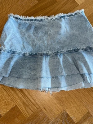 Ljusblå jeanskjol med volang - Trendig ljusblå jeanskjol med volangkant och råa kanter upptill och nedtill. Kjolen har en kort passform och är perfekt för dig som gillar en avslappnad och cool stil. Ingen synlig dragkedja eller knappar framtill.