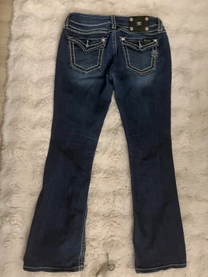 Mörkblå Miss Me jeans med kontrastsömmar - Säljer ett par mörkblå jeans från Miss Me med vita kontrastsömmar och broderade bakfickor. Jeansen har bootcut-modell och detaljerade ficklock med knappar. Perfekta för dig som gillar en snygg och markerad jeanslook.(⚠️OPS liten lila fläck på knät som inte går bort)