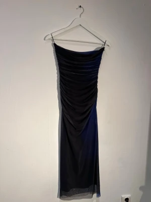 Mörkblå långklänning med drapering - Snygg mörkblå långklänning med smala axelband och draperad mesh över hela framsidan. Klänningen har en figurnära passform och är tillverkad i ett tunt, stretchigt material som ger en elegant siluett.