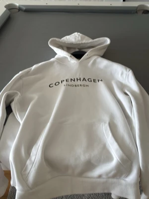 Vit hoodie från Lindbergh - Snygg vit hoodie från Lindbergh med trycket 'Copenhagen' på bröstet. Klassisk modell med huva och känguruficka framtill. Perfekt för en avslappnad och trendig stil. Mjukt material och ribbade muddar.