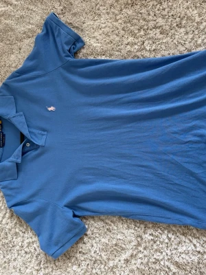 Blå pikétröja från Ralph Lauren - Klassisk blå pikétröja från Ralph Lauren med broderad logga på bröstet. Tröjan har krage, korta ärmar och knäppning framtill. Modellen är Classic Fit och tillverkad i mjuk bomull. Perfekt till sommaren 