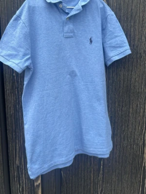 Blå pikétröja från Ralph Lauren - Klassisk ljusblå pikétröja från Ralph Lauren med korta ärmar och krage. Tröjan har knappar vid halsen och den ikoniska broderade loggan på bröstet. Perfekt för en avslappnad men ändå stilren look.