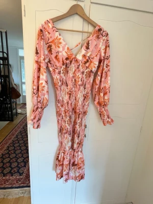 Blommig långklänning med volang - Romantisk stretchig långklänning i rosa och orange toner med blommigt mönster. Klänningen har v-ringning, långa ärmar med resår vid ärmslut och volang nertill. Perfekt för sommarens tillfällen.