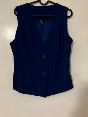 Kornblå väst med knappar - Stilren kornblå väst från H&M med två knappar framtill och justerbart band i ryggen. Djup V-ringning och figurnära passform. Perfekt för att lyfta en outfit med lite extra edge. Gjord i ett slätt tyg som känns bekvämt och snyggt.