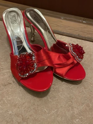 Röda pumps från Sergio Todzi med pärlor - Snygga röda pumps från Sergio Todzi med öppen tå och dekorativa pärlor framtill. Skorna har en glansig satinfinish och smal klack med extra bling. Perfekta för dig som vill sticka ut med färg och detaljer.