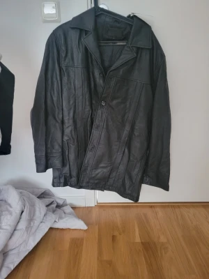 Svart lång skinnjacka med krage - Säljer en svart skinnjacka i längre modell med klassisk krage och knäppning framtill. Jackan har en stilren design med raka linjer och diskreta sömmar. Perfekt för dig som vill ha en tidlös och cool look.
