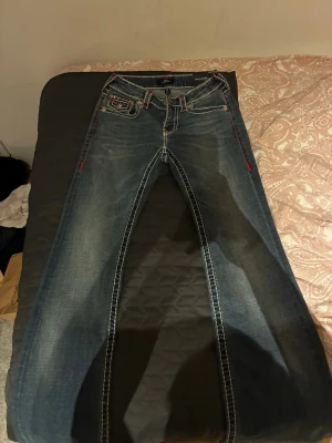  snygga True Religion  - Snygga mörkblå jeans från True Religion med kontrasterande vita sömmar och röda detaljer vid fickor och sidor. Klassisk femficksmodell med låg midja och raka ben. Perfekta för en cool och avslappnad stil.