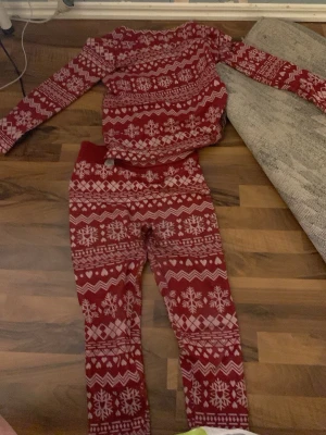 Röd julpyjamas med snöflingor - Mysig tvådelad pyjamas i rött med vita snöflingor och klassiskt julmönster. Setet består av långärmad tröja och byxor med resår i midjan. Perfekt för vinterkvällar och julmys.