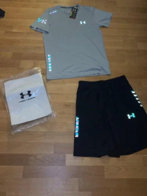 Under Armour  grå/svart - Snyggt träningsset från Under Armour med en ljusgrå t-shirt och svarta shorts. T-shirten har korta ärmar och tryckt logga på bröstet och ärmen. Shortsen har resår i midjan och logga på benet. Perfekt för gym eller löpning.