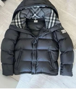 Svart pufferjacka från Burberry - Säljer en svart pufferjacka från Burberry med klassiskt rutigt foder i huvan. Jackan har dragkedja, knappar och två snedställda fickor framtill. Materialet är glansigt och quiltat för extra värme. Perfekt för kalla dagar.