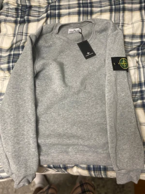 Grå sweatshirt från Stone Island - Snygg grå sweatshirt från Stone Island med klassisk rund hals och ikonisk patch på vänster ärm. Tröjan är tillverkad i mjukt bomullsmaterial och har ribbade muddar vid ärmslut och nederkant. Perfekt för en clean och stilren look.
