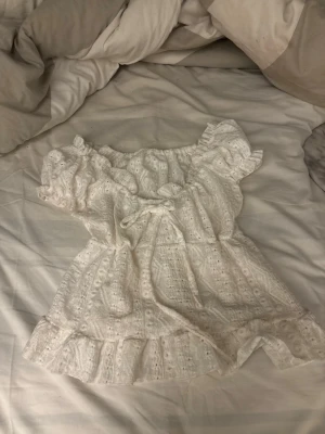 Vit broderad offshoulder-blus - Söt vit blus med broderade detaljer och volanger. Offshoulder-modell med korta puffärmar och resår vid axlarna. Tillverkad i ett lätt, luftigt tyg med romantisk känsla. Perfekt för en trendig och somrig look.