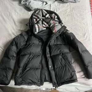 Burberry Jacka/Väst - Svart dunjacka med luva från Burberry London England, storlek M. Jackan har det klassiska Burberry-rutiga fodret i luvan och är perfekt för vår, höst och vinter. Den kan också göras om till en väst för extra flexibilitet. Notera att det finns lite problem vid kedjan på underarmen på höger arm.  