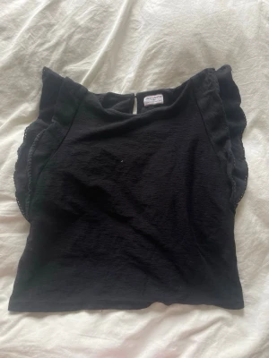 Svart topp med volang från Holly & White - Snygg svart topp från Holly & White med volangdetaljer längs axlarna. Toppen är ärmlös och har en rund halsringning samt en knapp i nacken. Perfekt att styla med jeans eller kjol för en trendig look.