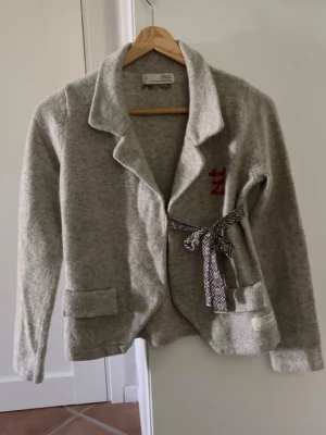 Grå kofta med knytband - Mysig grå kofta i ullmix med sjalkrage och dekorativt knytband i midjan. Koftan har två fickor framtill och en liten broderad detalj på bröstet. Perfekt för lager på lager och en avslappnad stil.