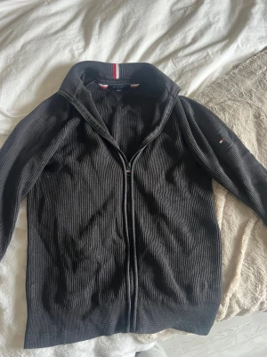 Svart ziptröja från Tommy Hilfiger - Snygg svart ziptröja från Tommy Hilfiger med ribbad struktur och ståkrage. Tröjan har dragkedja framtill och diskret logga på ärmen. Röd och vit randig detalj på kragen ger en sportig touch. Perfekt till en avslappnad stil.