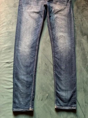 Blå Tiger of Sweden jeans - Klassiska blå jeans från Tiger of Sweden, modell Evolve. Snygg tvätt och diskreta slitningar. Jeansen har raka ben, fem fickor och detaljer i kopparfärg. Tvättas i 40 grader. 97 % bomull, 3 % elastan. W 31 Längd 34