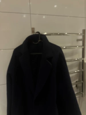 Mörkblå kappa från Gina Tricot - Stilren mörkblå kappa från Gina Tricot med klassisk krage och rak passform. Perfekt för dig som vill ha en enkel men snygg look. Kappan är i ett mjukt tyg som känns skönt att bära och passar till många olika outfits.