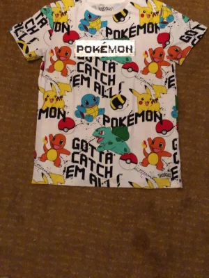 Pokemon t-shirt med färgglatt tryck - Cool t-shirt med Pokémon-motiv och texten 'Gotta catch em all!' över hela plagget. Pikachu, Squirtle, Charmander och Bulbasaur är med i designen. T-shirten är vit med färgglada detaljer och har korta ärmar. Perfekt för dig som älskar Pokémon!