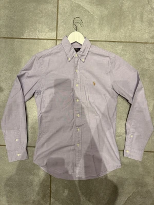 Ljuslila skjorta från Polo Ralph Lauren - Klassisk ljuslila skjorta från Polo Ralph Lauren med button-down krage och broderad logga på bröstet. Skjortan har långa ärmar, vita knappar och är tillverkad i bomull. Perfekt för en clean och stilren look. Passform slim fit.